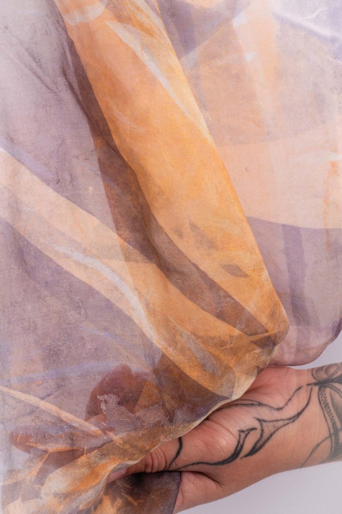 A sheer orange and aubergine patterned scarf draped over a tattooed hand, soft translucent fabric folds on display