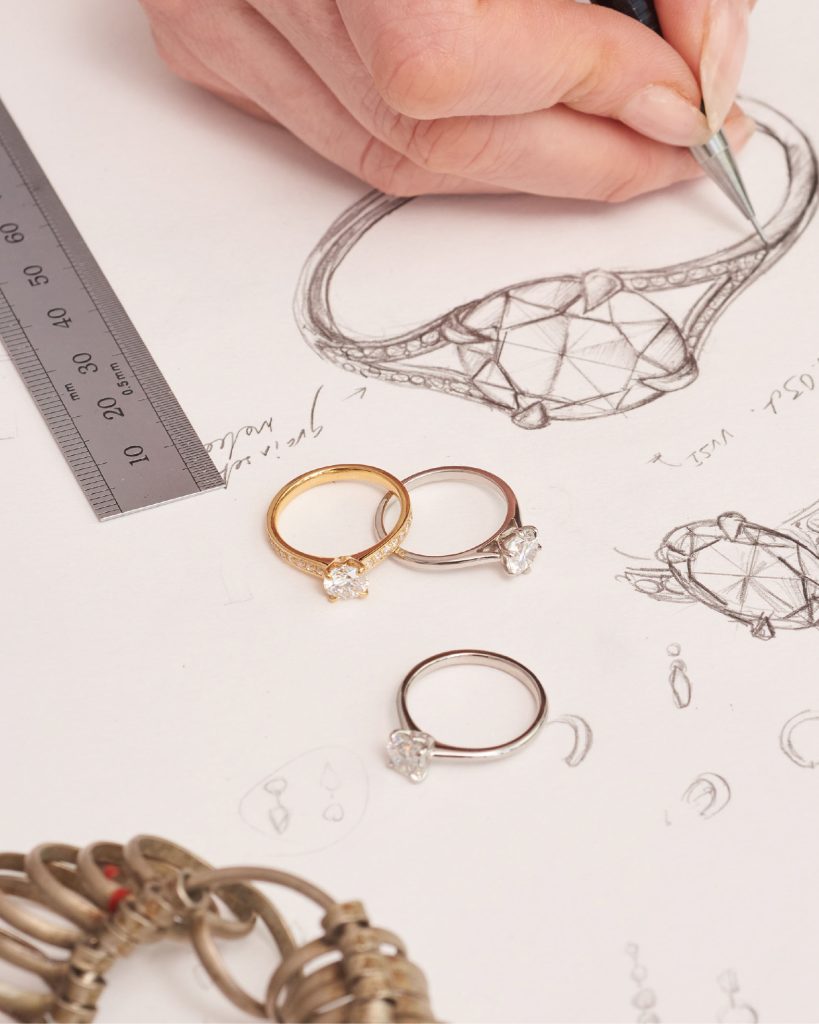 Jewellery designer sketching an intricate engagement ring beside three solitaire rings and a ruler on white paper