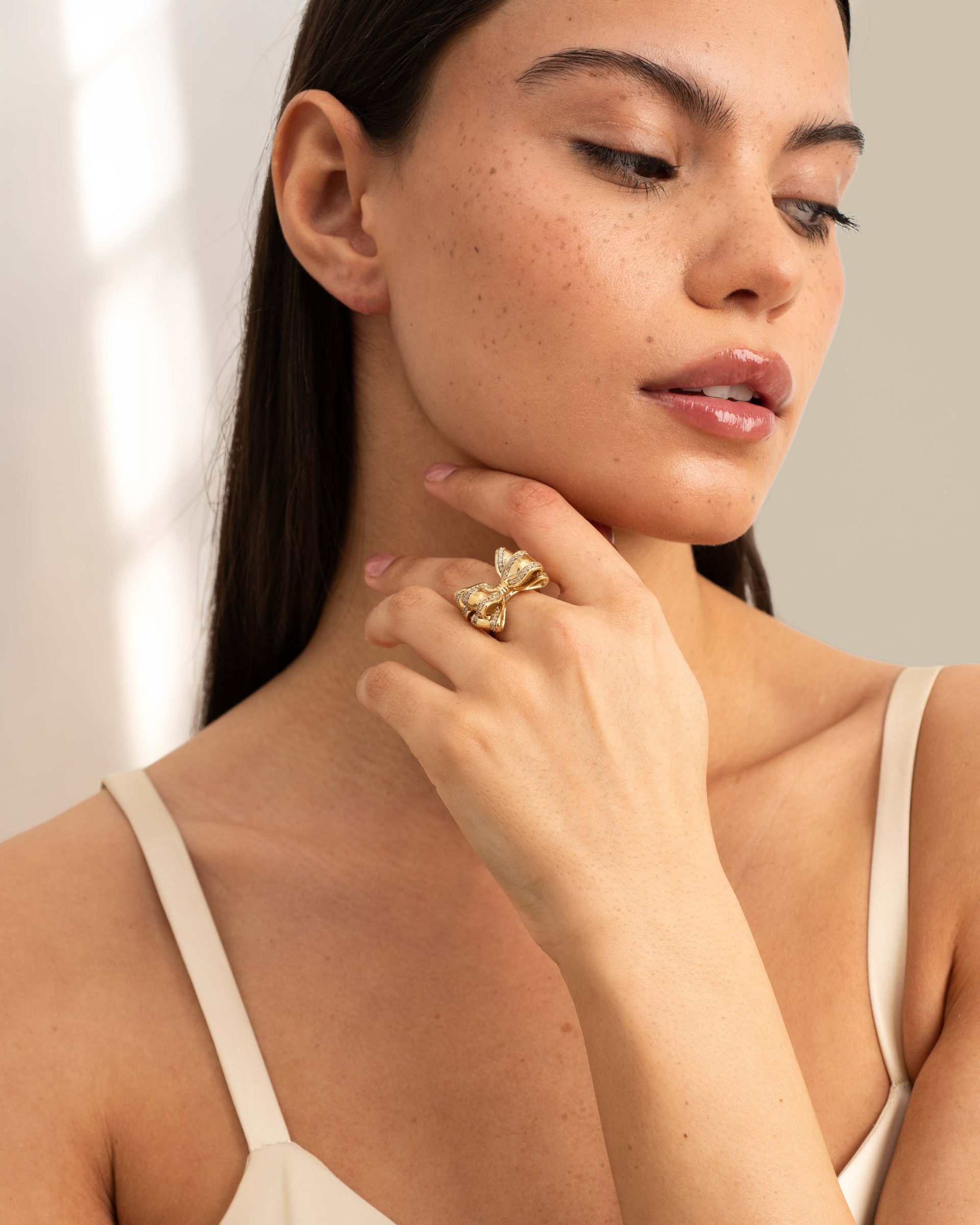 A close-up portrait shows a woman with glossy lips and freckles, wearing a sculptural gold ring in warm soft light.